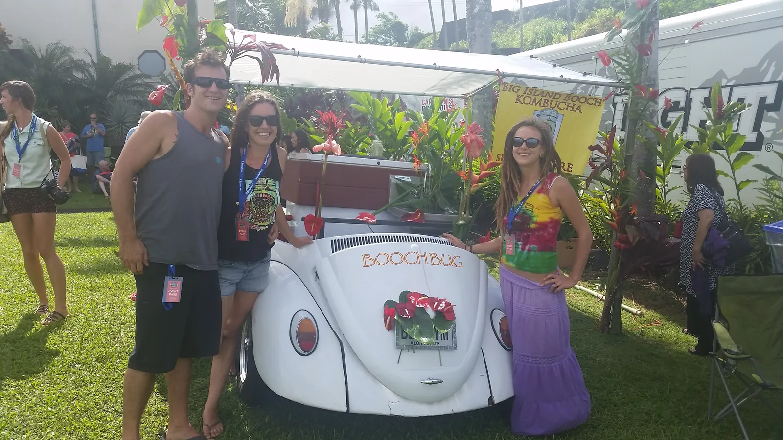 Festival-goers posing with the white Boochbug VW Beetle decorated with tropical flowers.