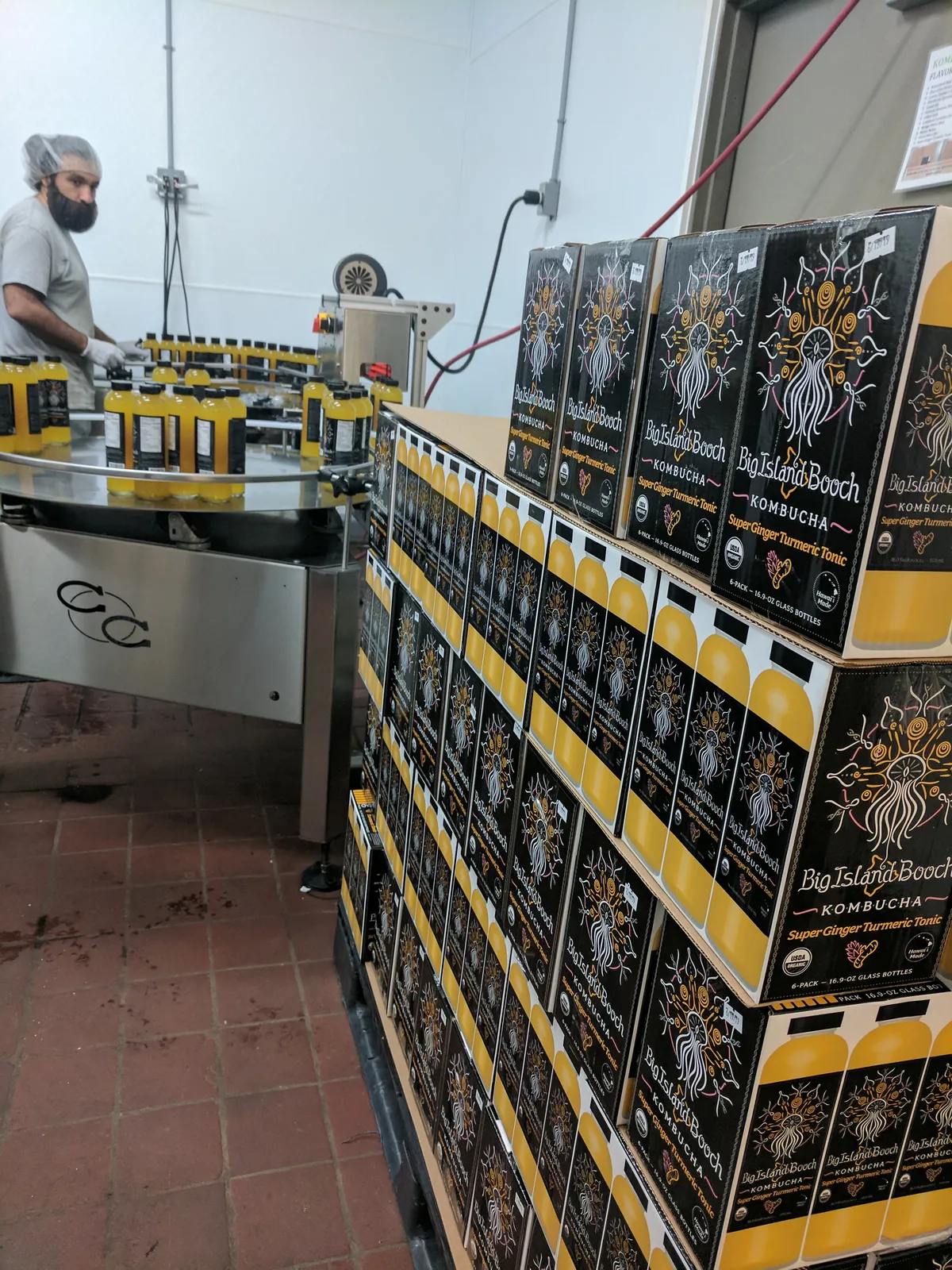 Stacked cases of Big Island Booch Super Ginger Turmeric Tonic beside the labeling machine.